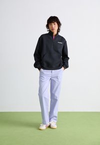 Sweatshirt noir zippé avec poche avant, associé à un pantalon lavande clair. Modèle portant des baskets blanches avec des accents roses.