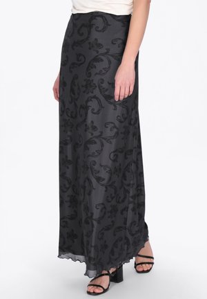 Kobieta ubrana w długą czarną spódnicę w kwiatowy wzór oraz czarne sandały na blokowym obcasie z paskami, stojąca na białym tle.