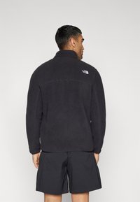 Svart fleecejakke med høy krage, teksturert stoff, og hvit logo på den øvre ryggen. Kombinert med svarte shorts.