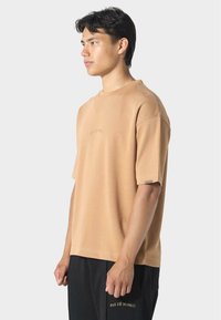 Beige bavlněné tričko s kulatým výstřihem, krátkými rukávy a jemně vyšitým logem na přední části. Mírně volný střih, měkká textura.