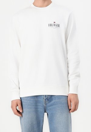 Mann trägt weißen Pullover mit langen Ärmeln mit dem Logo "Hilfiger New York" und hellblaue Jeans, steht vor einem weißen Hintergrund.