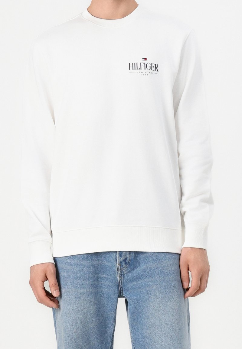 Homme portant un sweat-shirt blanc à manches longues avec le logo «Hilfiger New York» et un jean bleu clair, debout sur un fond blanc.