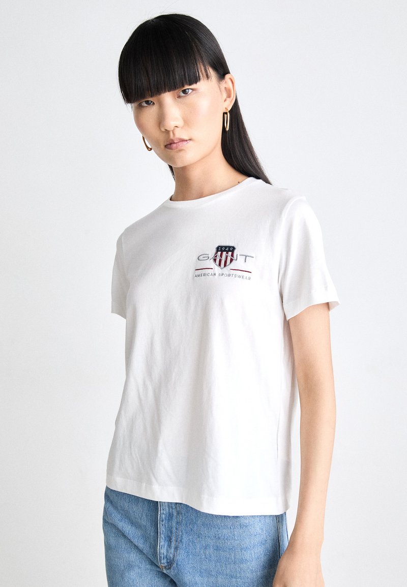T-shirt in cotone bianco, con un logo in blu navy e rosso. Collo tondo classico e maniche corte con un taglio rilassato. Indossato con jeans blu.