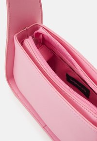 Růžová syntetická kabelka s hladkým povrchem, dvojitými zipovými přihrádkami a širokým klopovým uzávěrem. Značková etiketa viditelná uvnitř.