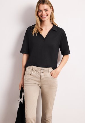 Jonge vrouw lacht, draagt een zwart kort mouw overhemd met kraag, beige broek en houdt een zwarte handtas vast tegen een effen achtergrond.