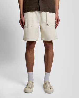 Man draagt beige korte broek met grote voorzakken, witte sokken en beige sneakers, staand tegen een grijze achtergrond.