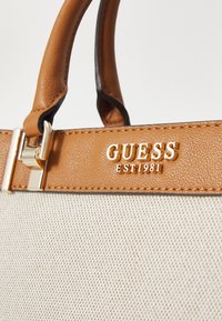 Manici in pelle marrone si collegano a un corpo in tela crema. Un accento in metallo color oro presenta il logo "GUESS" e la scritta "EST 1981".