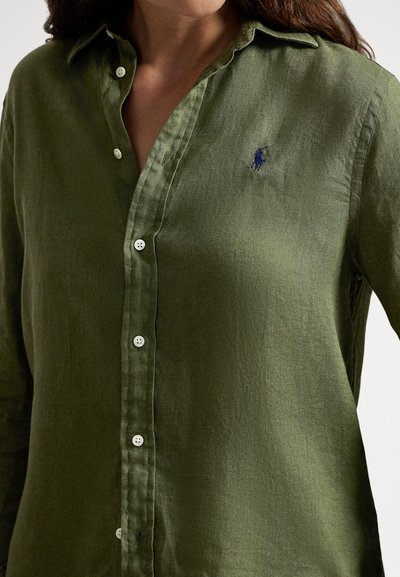 Olivzaļš lina krekls ar pogām, asu apkakli, baltām pogām un mazu zilu logotipu uz kreisās krūšu puses. Gluda tekstūra.