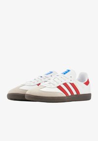 Bijele sportske tenisice s crvenim prugama, suđenim vrhom i smešnim gumenim đonom. Plavi jezičak s natpisom "Adidas Samba."
