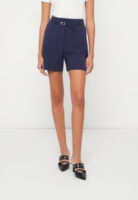 Tengerészkék, fazonra szabott sort, övvel, sima textúrával, oldalsó zsebekkel és enyhén laza fazonnal. Fekete magassarkú mules-szal párosítva.