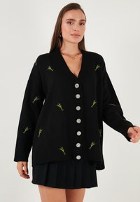 Crni kardigan s V-izrezom, oversized krojem, cvjetnim vezom u zelenoj i žutoj boji, te srebrnim dekorativnim gumbima duž prednje strane.