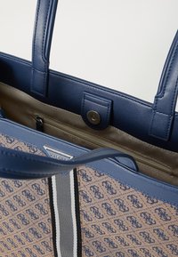 Sac fourre-tout bleu marine avec un corps en toile présentant un motif de logo répété. Accents en cuir et poche intérieure zippée. Détail rayé sur le côté.