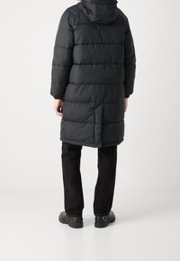 Person trägt einen langen schwarzen Steppmantel mit Kapuze, schwarze Hose und schwarze Schuhe und steht vor einem schlichten hellen Hintergrund.