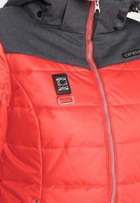 Veste matelassée rouge et gris foncé avec fermeture éclair devant, insigne métallique et détail boucle en tissu sur la poitrine.