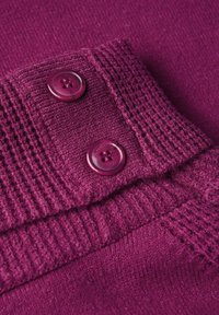 Fialová pletenina se dvěma lesklými knoflíky. Textura obsahuje žebrované detaily blízko knoflíků, které zdůrazňují strukturu oděvu.