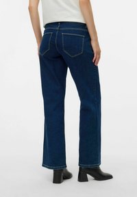 Tumši zili džinsu bikses ar taisnu kāju, kurām ir divi aizmugurējie kabati un dzelteni šuvuma akcenti, saskaņotas ar melnajām apaviem ar papēžiem.