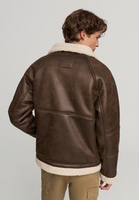 Brązowa skórzana kurtka z kremowym kołnierzem z sztucznego barana, charakteryzująca się gładką fakturą i starannym przeszyciem wzdłuż pleców i rękawów.