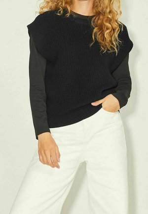 Kobieta o kręconych włosach, ubrana w czarną, bezrękawnikową dzianinową kamizelkę na czarnym długim rękawie i białe spodnie o szerokich nogawkach, z ręką w kieszeni.