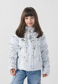 Chaqueta blanca con lentejuelas y cuello alto, con tela texturizada y diseño acolchado. Combinada con vaqueros azules, mostrando un look casual.