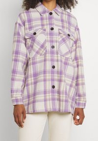 Kvinna klädd i en långärmad skjorta med knappar i lila och beige rutmönster, två bröstfickor, tillsammans med off-white byxor.