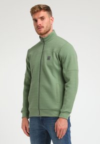 Chaqueta de forro polar verde con textura acolchada, cuello alto y bolsillos laterales. Presenta un pequeño logo en el pecho. Diseñada para mantener el calor.