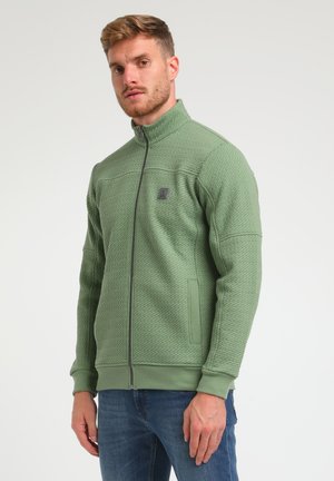 Chaqueta de forro polar verde con textura acolchada, cuello alto y bolsillos laterales. Presenta un pequeño logo en el pecho. Diseñada para mantener el calor.