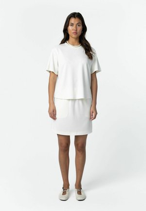 Vrouw staand, gekleed in een wit kortermig shirt, een rok tot op de knie, een parelketting, oorringen in ringvorm en witte veter schoenen, op een witte achtergrond.