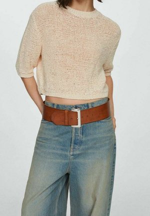 Henkilö, jolla on päällään kerma värinen neulottu lyhyt pusero, korkeavyötäröiset haalistuneet siniset farkut ja leveä ruskea nahkavyö hopeisella soljella.