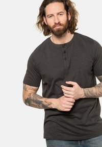 Tmavě šedé tričko henley s krátkým rukávem, které má tři knoflíky u límce, vyrobené z měkkého materiálu. Model má viditelné tetování na pažích.