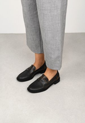 Mocasines de cuero negro con un acabado suave y detalle de banda texturizada. Suela de perfil bajo y acentos naranjas en contraste. Combinados con pantalones cropped grises.