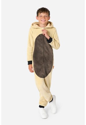 Junge lächelt und steht, trägt einen beige-braunen Kapuzen-Overall mit schwarzen Bündchen und weißen Turnschuhen vor einfarbigem Hintergrund.