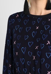 Schwarzes Stoffoberteil mit einem Muster aus blauen und rosa Herzen und Kreuzen, rundem Ausschnitt und langen Ärmeln. Die Textur wirkt glatt.