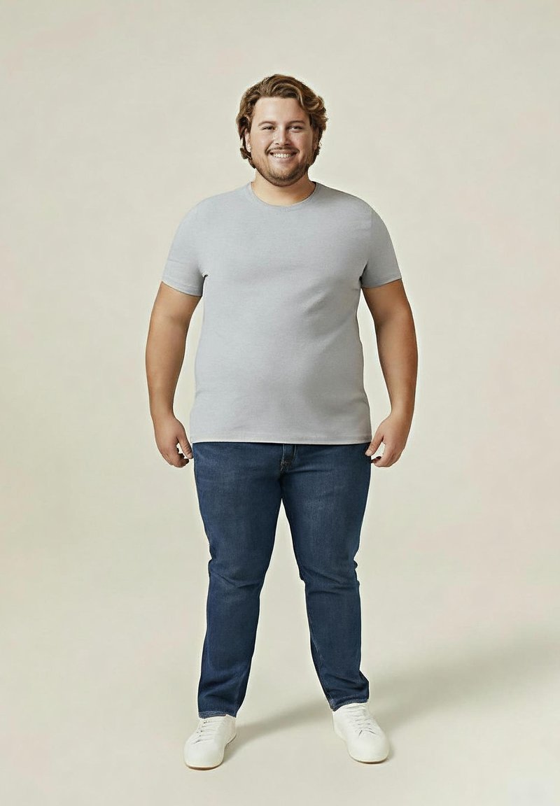 Lächelnder Mann mit hellbraunen Haaren, der ein graues T-Shirt, blaue Jeans und weiße Sneaker trägt, steht vor einem schlichten beigen Hintergrund.