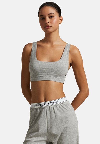 Kobieta w szarym krótkim topie na ramiączkach i pasujących szarych spodniach do noszenia w domu z białym paskiem Polo Ralph Lauren, stojąca na białym tle.