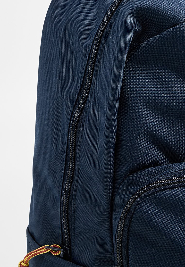 Sac à dos bleu marine avec un tissu texturé, deux compartiments zippés et un détail de sangle tissée avec des accents jaunes et rouges.
