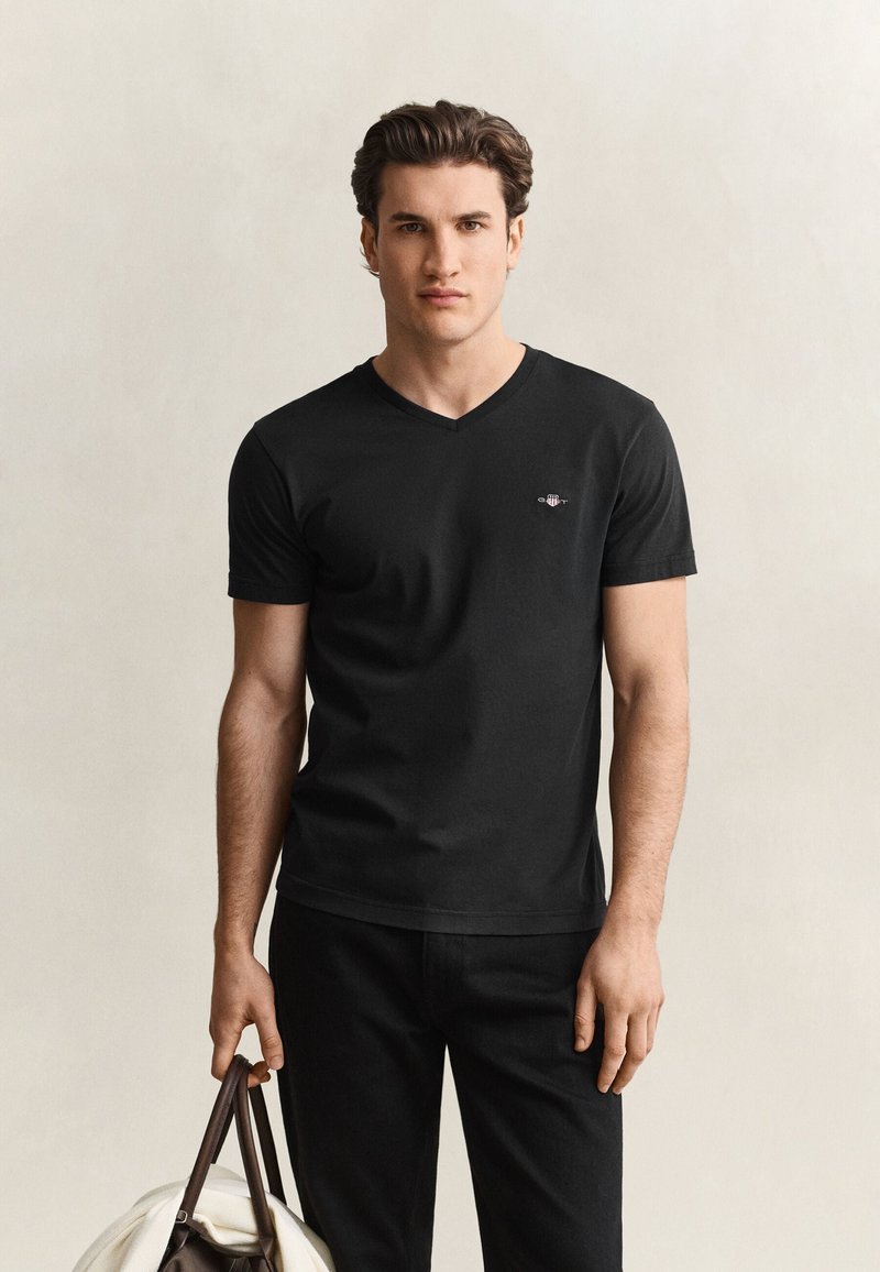 Ung mand med brunt hår iført sort V-hals T-shirt og bukser, holder en beige og brun sportstaske mod en ensfarvet baggrund.