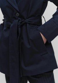 Granatowy płaszcz z paskiem o dopasowanym kroju, z pojedynczym zapięciem na guzik i kieszeniami po bokach. Miękki materiał o gładkiej strukturze.