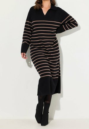Femme portant une robe midi noire avec des rayures horizontales marron et un col, associée à des bottines noires, debout devant un fond blanc.
