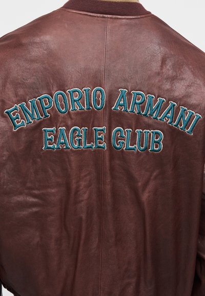 Καφέ δερμάτινο μπουφάν μπομπέρ με κεντημένο το λογότυπο "EMPORIO ARMANI EAGLE CLUB" σε τιρκουάζ στη ράχη. Ομαλή υφή, με ριμπ γιακά και μανσέτες.