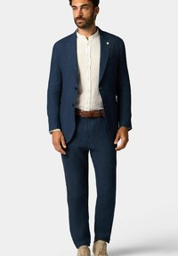 Completo in lino blu navy con giacca su misura, pantaloni coordinati e una camicia in lino di colore chiaro. Presenta una cintura intrecciata marrone testurizzata e scarpe casual.