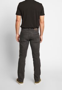 Vaqueros de mezclilla gris oscuro, con corte recto, cinco bolsillos y una parche de cuero marrón en la cintura. Vista desde atrás.