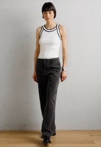 Débardeur blanc à rayures avec des accents noirs, associé à un pantalon gris foncé taille haute. Modèle portant des chaussures noires, debout sur un sol en bois.