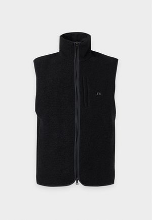 Man die een zwart vest met rits draagt over een donkergroene en zwarte gebreide trui, zwarte broek en zwarte sneakers, staand met handen in de zakken.