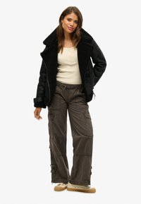 Schwarze Schaffelljacke über einem crèmefarbenen Henley-Shirt, kombiniert mit locker sitzenden grauen Cargo-Hosen und crèmefarbenen Sneakers, die seitliche Taschen und Reißverschlüsse aufweisen.