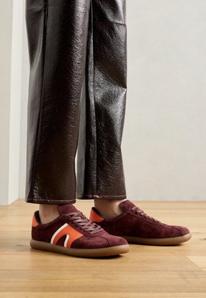 Burgundowe i pomarańczowe zamszowe trampki z białym akcentem, z okrągłym noskiem i brązową gumową podeszwą, w zestawie z błyszczącymi brązowymi spodniami.