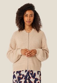 Béžový pletený cardigan s texturovaným vzorem, se zipovým zapínáním a vroubkovanými manžetami. Nosený přes šaty s květinovým potiskem, které mají tmavě modré a oranžové akcenty.