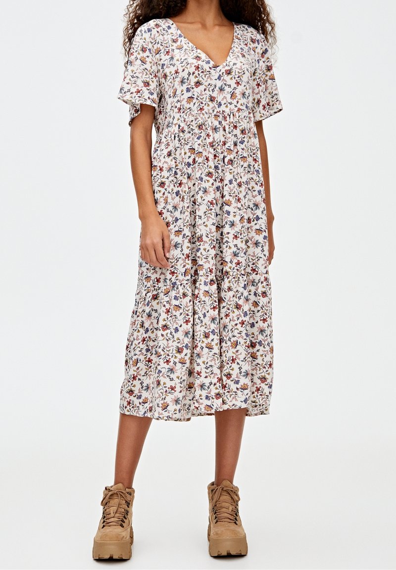 Femme portant une robe midi blanche à fleurs avec manches courtes et col en V, associée à des bottines marron clair à lacets.