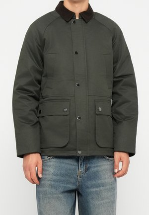 Uomo che indossa una giacca verde scuro con bottoni, due tasche frontali con pattina e un colletto nero, abbinata a jeans blu.