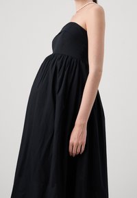 Robe noire sans bretelles avec un corsage ajusté et une jupe pleine froncée. Texture de tissu lisse, longueur au genou, portée avec un collier de perles.