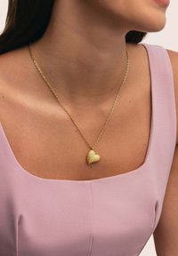 Goldene Herzanhänger-Halskette mit strukturiertem Oberflächen geh�ängt an einer feinen Kette, getragen über einem hellrosa ärmellosen Oberteil mit quadratischem Ausschnitt.